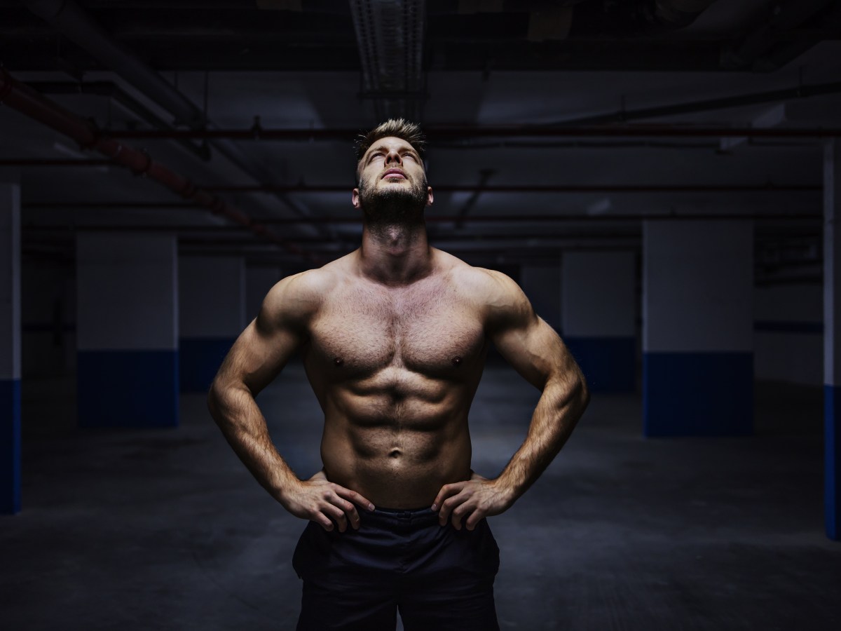 Powerful shirtless muscular sportsman standing in garage at night with hands on hips and looking up. Urban life.