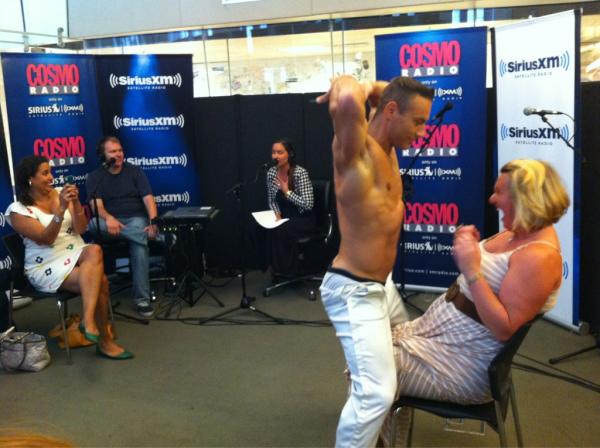 CosmoRadio Shirtless dancer performs close to seated woman in radio studio setting.