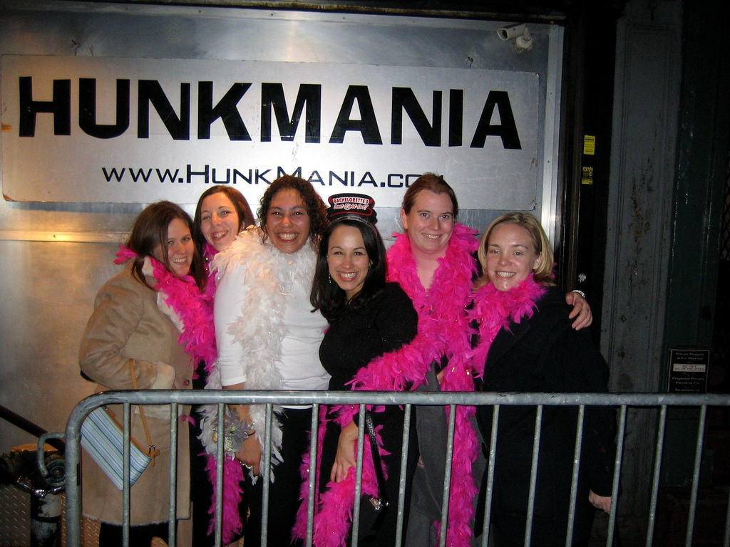 HunkmaniaSign Group of six women smiling, wearing feather boas, in front of 'Hunkmania' sign.