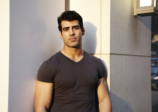 Man in a dark t-shirt leans against a wall, with soft outdoor lighting.
